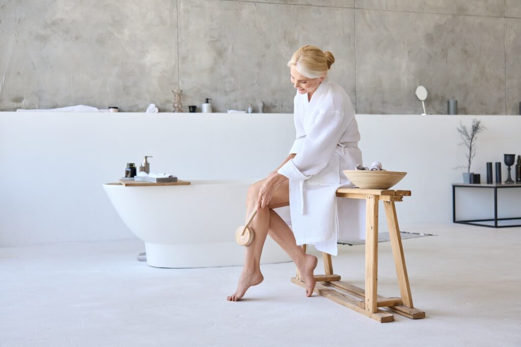 Middle aged mature woman in robe massaging legs sitting in modern bathroom.